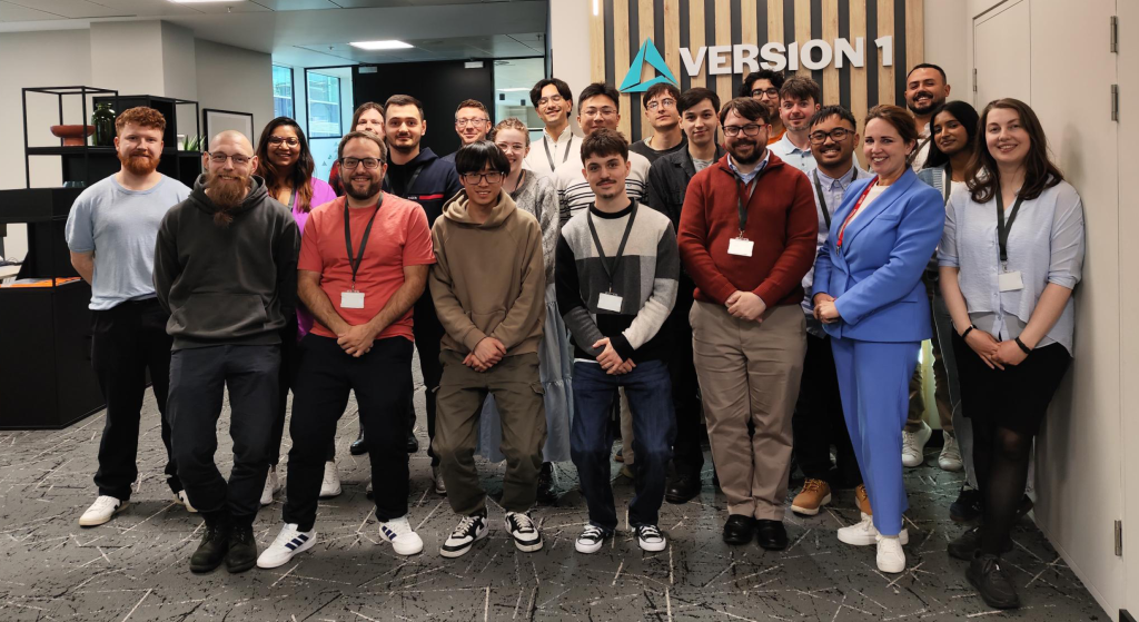 A large group of early‑career staff members stand together in a modern office space, facing the camera. The group is arranged in rows and wearing casual to business-casual clothing, many with lanyards and ID badges. Behind the group, a wooden feature wall displays the “VERSION 1” logo and branding. The office includes shelving units, a glass-walled room, and overhead lighting, creating a professional workplace setting.