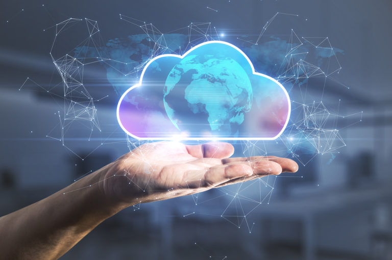A close-up of a human hand held open with a glowing cloud icon hovering above the palm. Inside the cloud shape is a semi-transparent globe showing continents, symbolising global cloud computing. The background features a blurred office-like setting with a network of light lines and nodes, representing digital connectivity and data flow. The overall colour palette includes cool tones of blue and purple with bright highlights around the cloud.