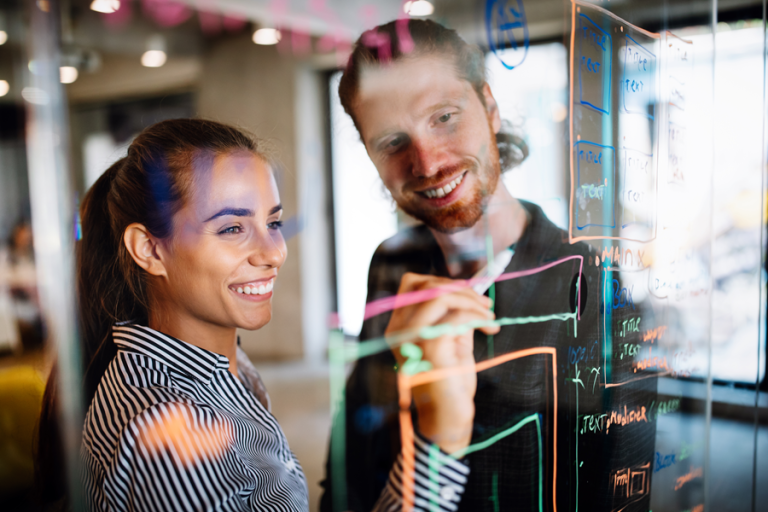 Two colleagues collaborating at a glass whiteboard, reviewing and mapping ideas during a strategy and innovation workshop.
