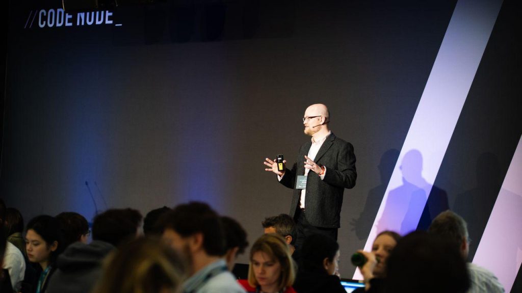 Man presenting on stage at a conference, holding a device and speaking to a seated audience.