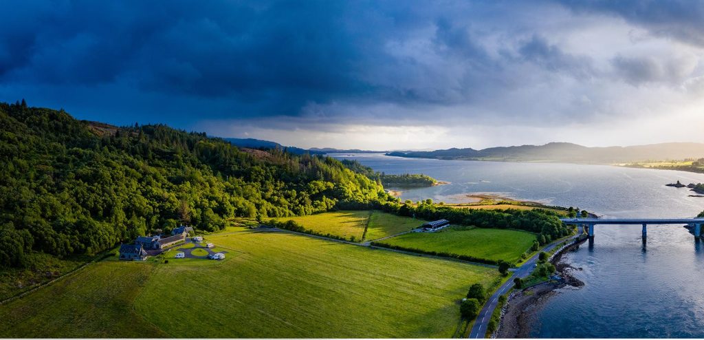 A wide, panoramic landscape view showing a coastal countryside with rolling green fields, dense woodland, and a large body of water stretching into the distance. In the foreground, a rural property with several buildings sits within open farmland bordered by hedgerows and narrow roads. To the right, a long bridge extends across the water, connecting land on either side. The shoreline curves gently around the bay, with patches of trees and small inlets visible. In the background, low hills and mountains fade into the horizon. The sky is dramatic, with dark storm clouds on the left gradually giving way to brighter light on the right, casting a mix of shadow and sunlight across the scene.