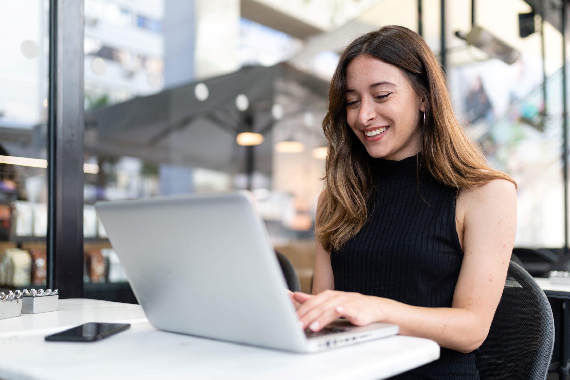 The image shows an indoor café or modern workspace setting with a single person seated at a table using a laptop. The person is positioned in the foreground, facing slightly to the side, with both hands on the keyboard. The laptop is open on a light-colored table, and a smartphone lies nearby. The background is softly blurred but suggests a contemporary environment with large windows, natural daylight, and hanging lights. There are structural elements and possibly shelves or displays visible, contributing to a relaxed yet professional atmosphere. The scene highlights focused computer use in a casual, well-lit environment that blends elements of work and leisure.
