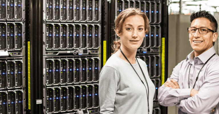 A data center environment is shown with rows of server racks filling the background. The racks contain multiple stacked servers with uniform rectangular units, each displaying small indicator lights in blue and green, suggesting active systems. The equipment is arranged in tall, dense columns, creating a structured and highly technical setting. In the foreground, two individuals stand in front of the server racks. Both individuals wear lanyards with ID badges, indicating a professional or secured workplace environment. One individual stands slightly in front, facing toward the camera, while the other stands to the side with arms crossed. Both are positioned close to the equipment, suggesting involvement in monitoring, maintenance, or management of the infrastructure. The lighting is bright and even, highlighting the metallic surfaces of the servers and the clean, organized layout of the facility. The color palette is dominated by dark tones from the server units, contrasted by the lighter clothing of the individuals and the small, bright LED indicators. Overall, the image conveys a professional IT or data infrastructure setting focused on server management and technical operations.