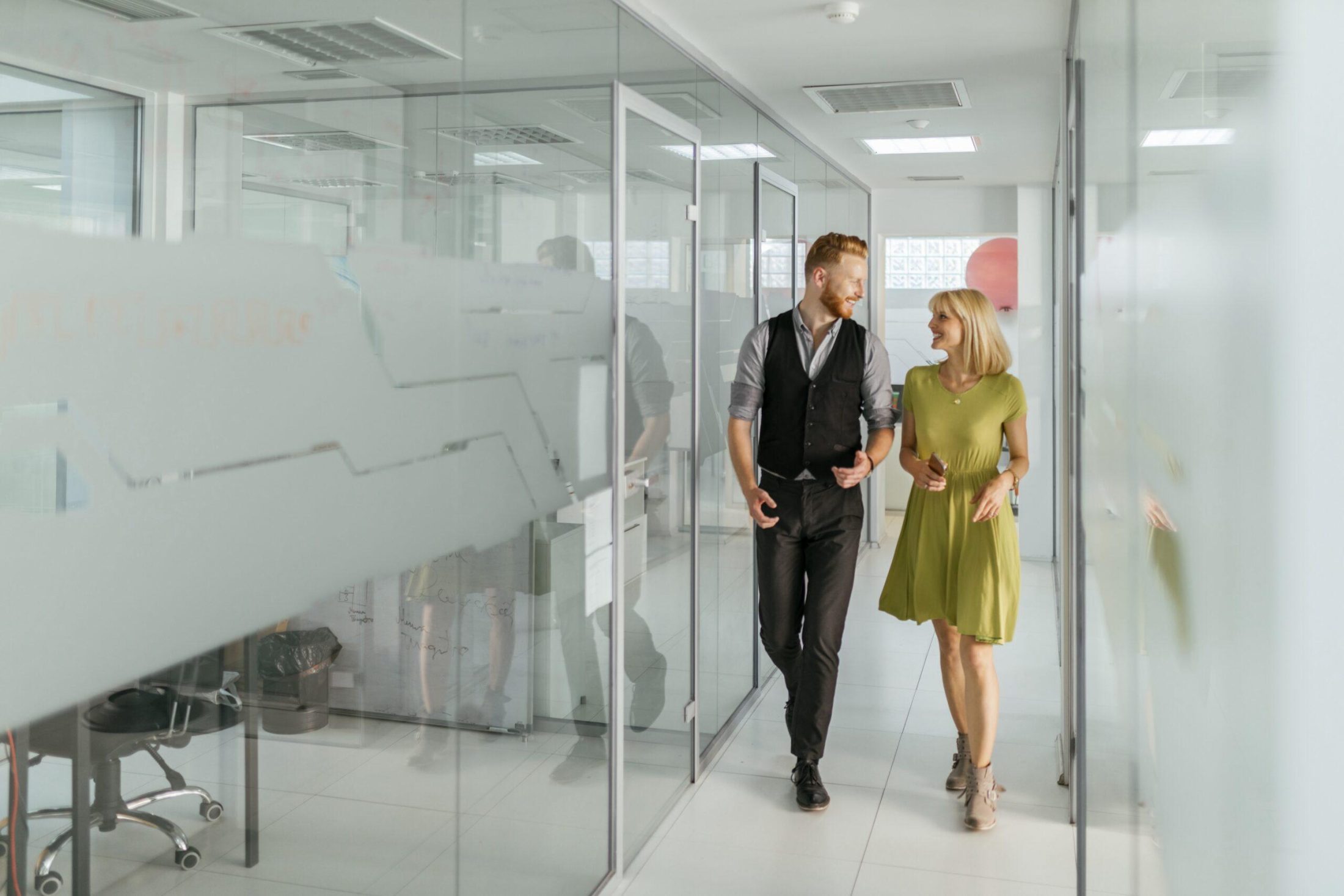 A bright, modern office corridor with glass-walled meeting rooms on both sides. Two people are walking side by side down the hallway, appearing to be engaged in conversation. One individual is wearing a dark vest over a long-sleeve shirt with dark trousers and dress shoes, while the other is wearing a light green short-sleeve dress paired with ankle boots. Both are mid-step, facing slightly toward each other. The glass walls have frosted sections and reflect parts of the corridor and the people. Overhead, rectangular ceiling lights evenly illuminate the clean, white space. In the background, an open door and a wall with a circular pink design element are visible.
