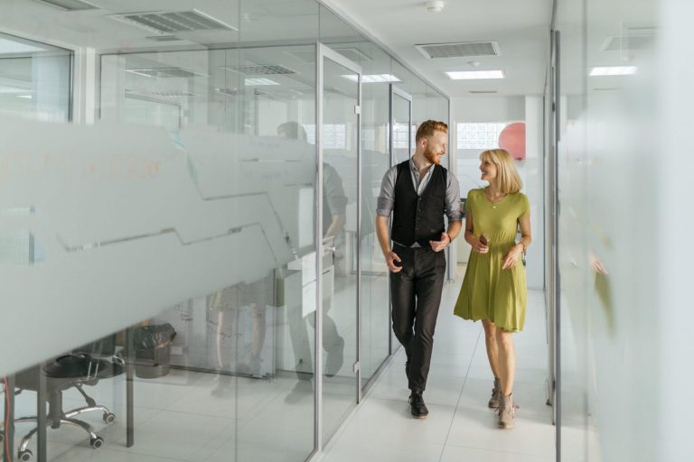 A bright, modern office corridor with glass-walled meeting rooms on both sides. Two people are walking side by side down the hallway, appearing to be engaged in conversation. One individual is wearing a dark vest over a long-sleeve shirt with dark trousers and dress shoes, while the other is wearing a light green short-sleeve dress paired with ankle boots. Both are mid-step, facing slightly toward each other. The glass walls have frosted sections and reflect parts of the corridor and the people. Overhead, rectangular ceiling lights evenly illuminate the clean, white space. In the background, an open door and a wall with a circular pink design element are visible.