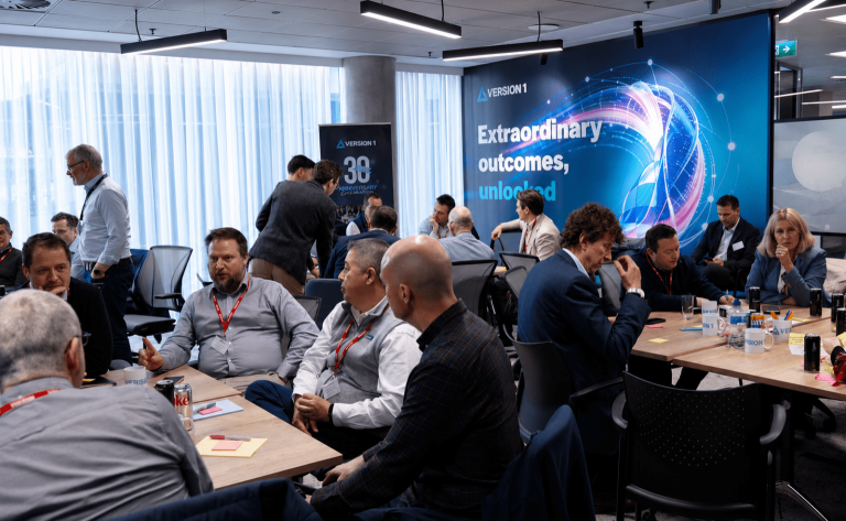 Business workshop with professionals seated in groups discussing ideas in a modern office, with a large “Extraordinary outcomes, unlocked” presentation backdrop.