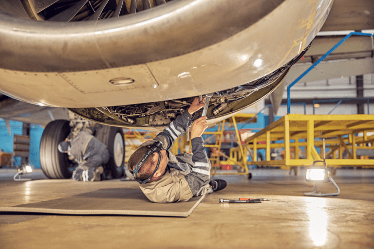 A maintenance technician lies on a mat beneath a large aircraft engine inside an aircraft hangar, using a wrench to work on exposed mechanical components. Another technician works near the landing gear in the background, while maintenance platforms, tools, and portable work lights surround the aircraft. The hangar is brightly lit, highlighting the underside of the plane and the engine housing.