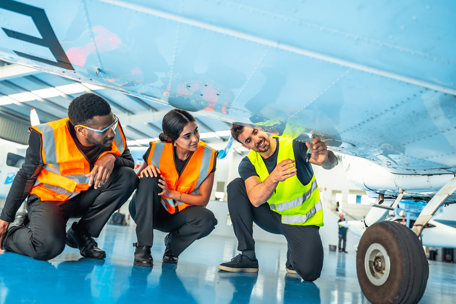 Three aircraft maintenance trainees wearing high-visibility safety vests and protective eyewear crouch beneath the wing of an aircraft inside a hangar. One trainee points to an inspection panel on the underside of the wing while the others observe closely. The aircraft’s landing gear wheel is visible on the right, and the brightly lit hangar features a blue floor and additional aircraft in the background.