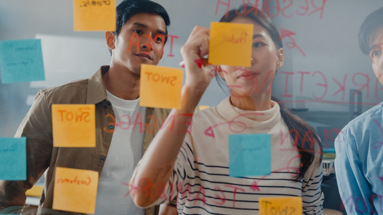 Team members collaborating in a workshop, placing sticky notes on a glass board while mapping ideas and priorities for a service design project.