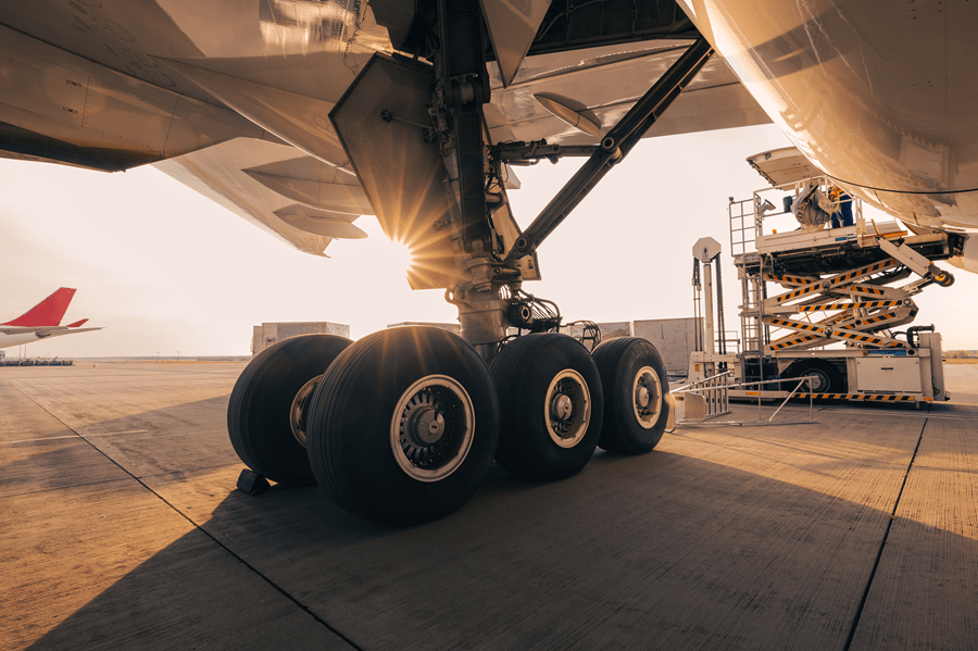 Close-up view of a large aircraft’s main landing gear on an airport apron at sunset. Multiple wheels and the landing gear assembly are visible beneath the aircraft, illuminated by warm sunlight creating a starburst effect. A maintenance platform lift is positioned beside the aircraft for servicing, while another aircraft is visible in the distance on the left side of the frame.