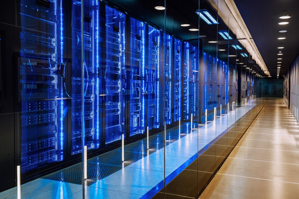 Modern data center server room with rows of glass-enclosed racks illuminated by blue LED lights, extending down a long, clean corridor.