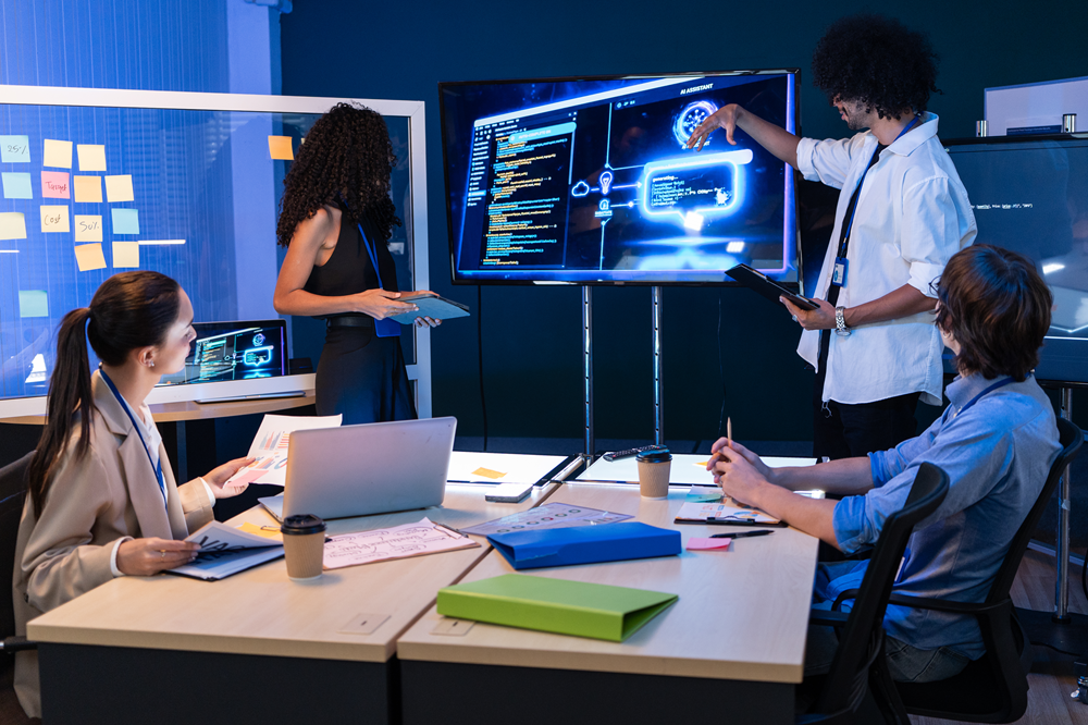 A group of four people in a modern office meeting room collaborate around a large digital display showing software code, cloud computing graphics, and technology diagrams. One person stands at the screen presenting with a tablet while three others sit at a conference table with laptops, notebooks, and office supplies. Sticky notes are visible on a glass wall in the background, suggesting a planning or development session. The room is lit with cool blue tones that emphasize the technology-focused environment.