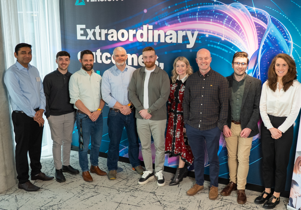 Group photo of nine people standing indoors in front of a large, colorful display wall featuring the visible text “Extraordinary outcomes.” The display has a blue background with sweeping pink, purple, and white light-like graphical elements. The people are arranged in a line facing the camera and wearing a mix of business and smart-casual clothing, including shirts, jackets, trousers, dresses, and boots. The setting appears to be a corporate or professional event space, with a patterned light-colored floor and white curtains along the left side of the image. Bright, even lighting illuminates the group and backdrop.