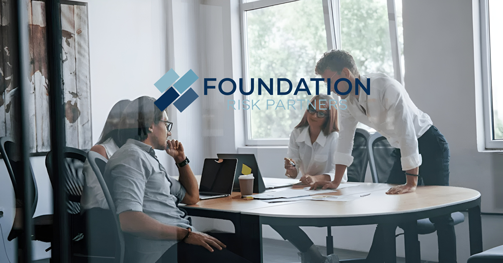 Foundation Risk Partners logo overlaid on a modern office meeting scene. Three people are gathered around a round conference table with laptops, papers, and a takeaway coffee cup. Large windows provide bright natural light in the background. The logo features a blue geometric icon and the text “FOUNDATION RISK PARTNERS” centered across the image.