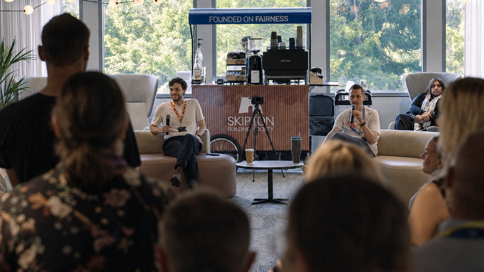 Audience members sit facing a panel discussion in a bright lounge-style event space. Two speakers are seated at the front near a small table and a branded coffee station displaying the text “SKIPTON Building Society” and “Founded on Fairness.” Large windows provide natural light, and a camera on a tripod is positioned between the speakers and the audience.
