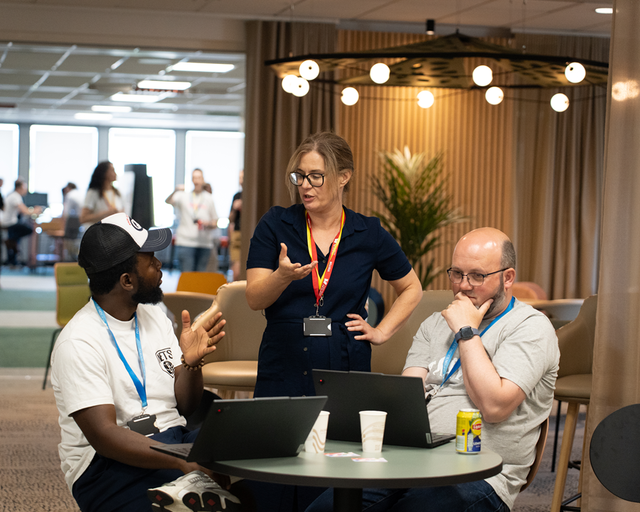 Three people collaborate around a small table in a modern office lounge area. Two participants sit with open laptops while a standing participant gestures during the discussion. Coffee cups and a canned drink sit on the table, and soft lighting, plants, and seating areas are visible in the background.