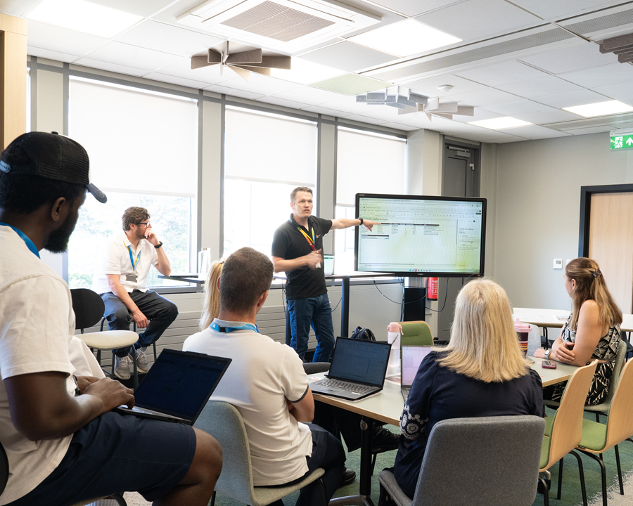 A group of people sit around tables in a modern meeting room while one presenter stands beside a large digital display, pointing to a spreadsheet during a workshop or team training session. Several attendees use laptops as they follow the presentation, with large windows providing natural light in the background.