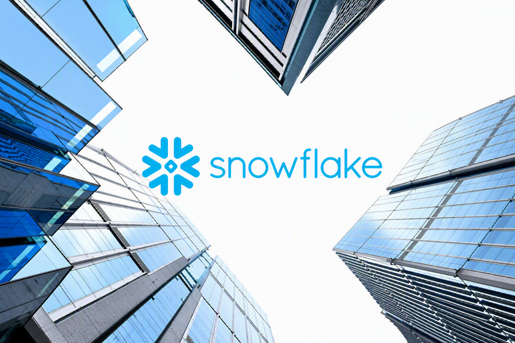 Snowflake logo centered against a bright sky, surrounded by four modern glass office towers viewed from below. The reflective blue-and-silver buildings frame the image from each corner, creating a clean, symmetrical composition that represents cloud data platforms, enterprise technology, and digital innovation.