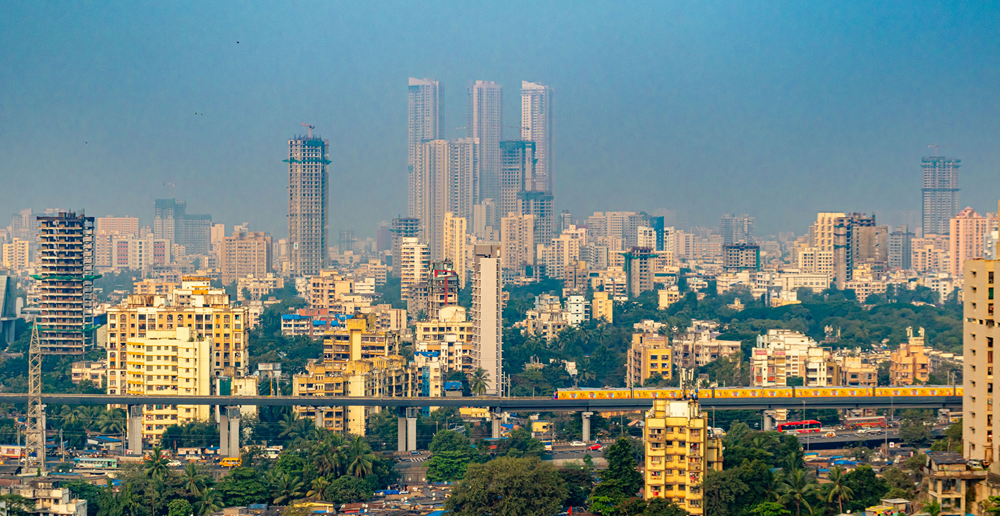 Panoramic view of Pune urban skyline featuring a dense cluster of high-rise residential and commercial buildings. An elevated metro line runs across the foreground with a yellow train in motion, while pockets of greenery and lower-rise structures sit beneath the towering skyscrapers. The scene is captured under a hazy blue sky, highlighting the scale and density of the city.